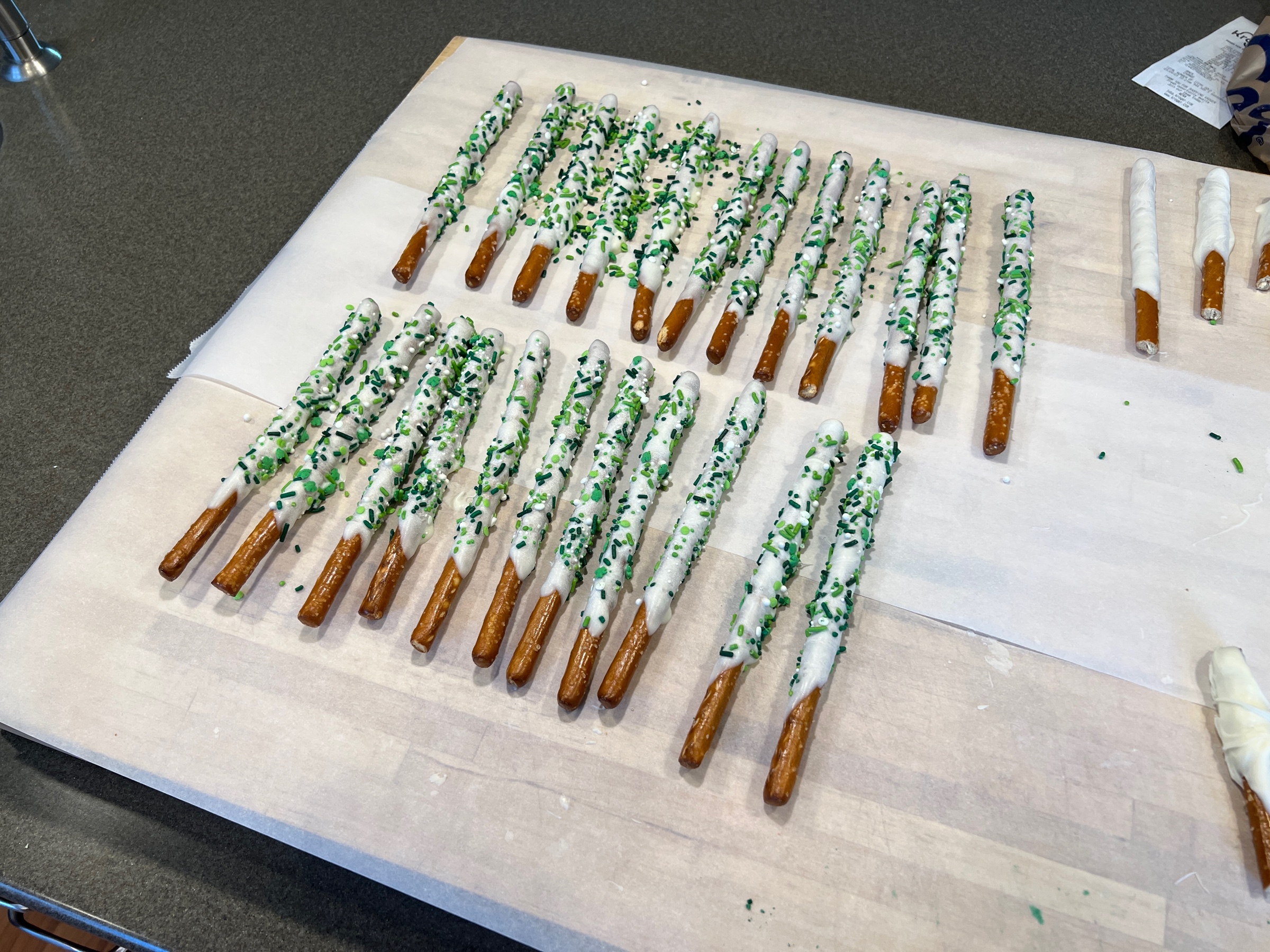 Pretzel rods, dipped in white chocolate and decorated with green sprinkles, cooling on parchment paper. At the edge of the photo are a few broken pretzel rods, dipped in white chocolate, but undecorated.
