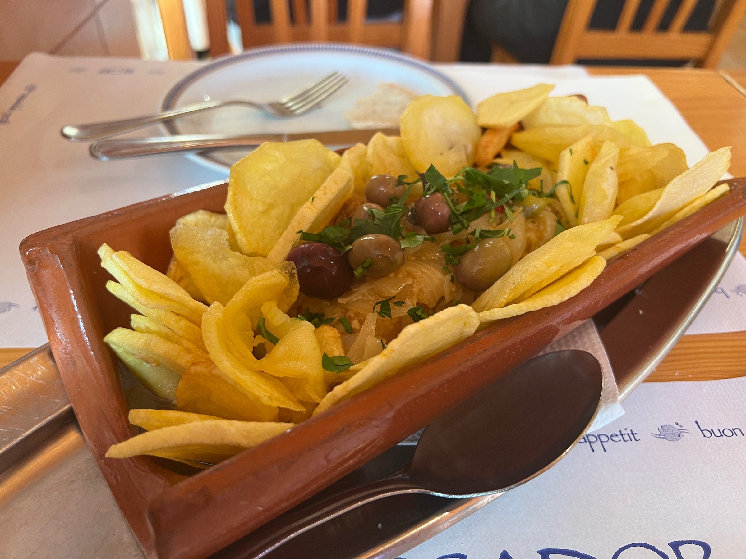 A huge portion of braised salted cod with onions and olives, presented in a bed of chips, and served in a red clay roofing tile.