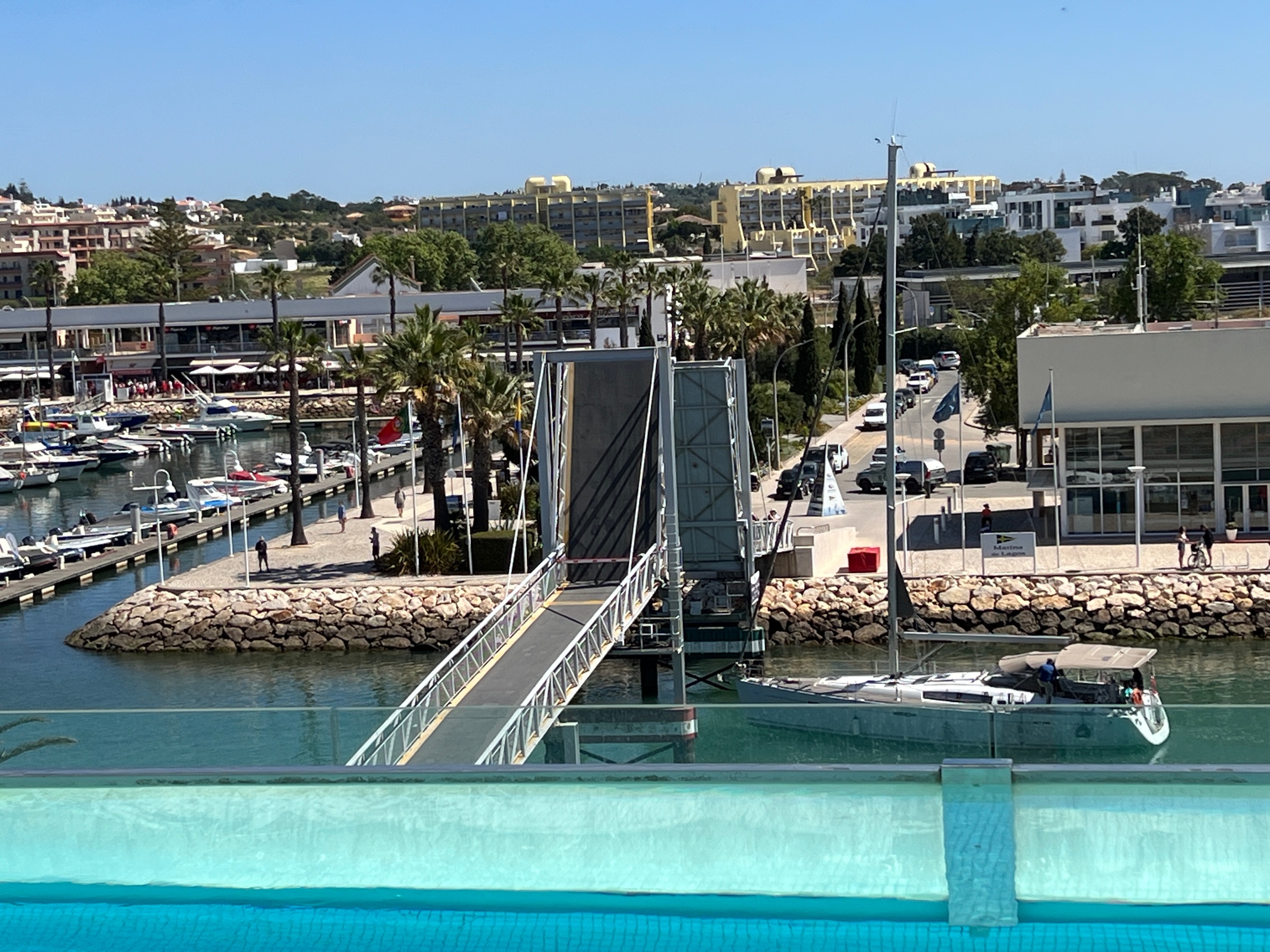 a pedestrian drawbridge at the mouth of the marina that has been raised to allow a masted yacht to pass