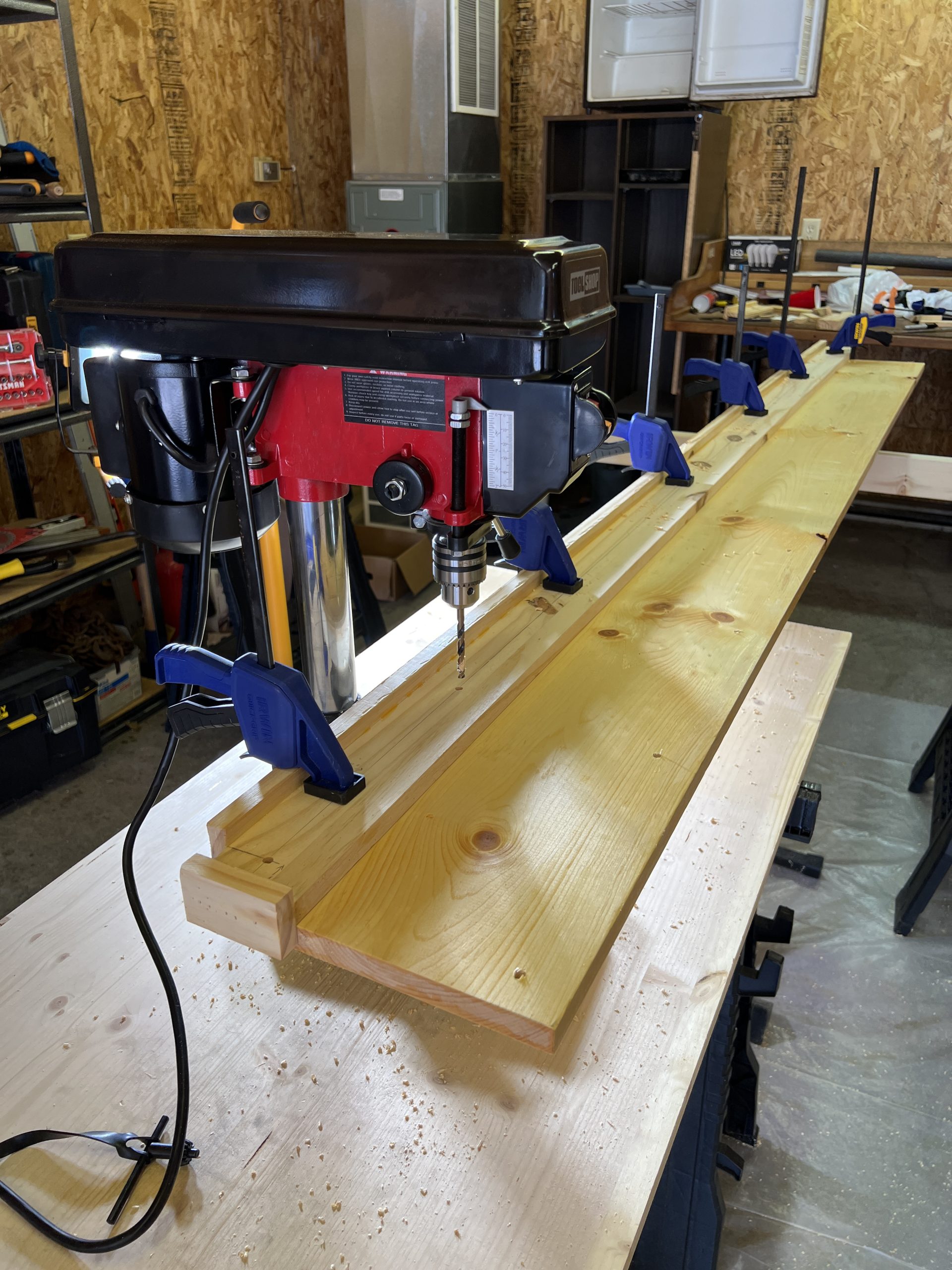 A 86 inch 1x10 laying on the table of a small drill press, with a long jig clamped to it.