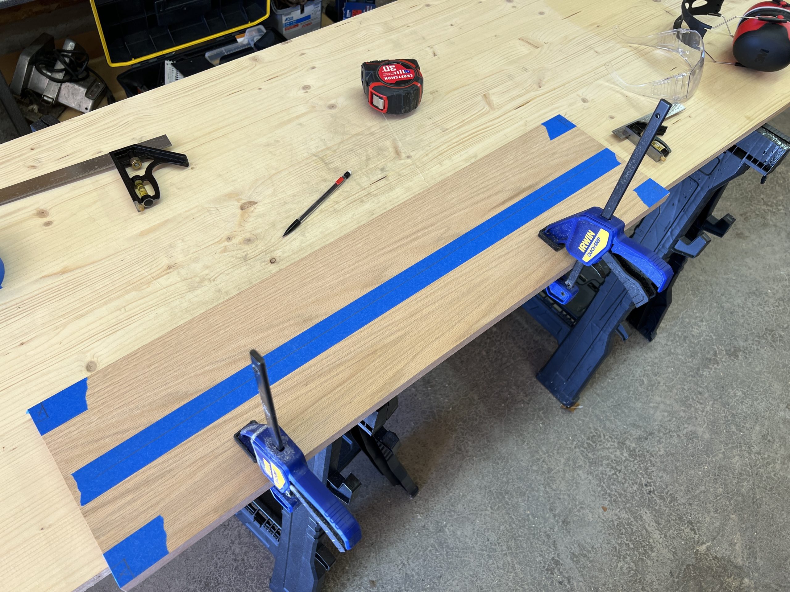 An oak board clamped to a woodworking bench. Painter’s tape covers the corners and midline where measurements are marked.